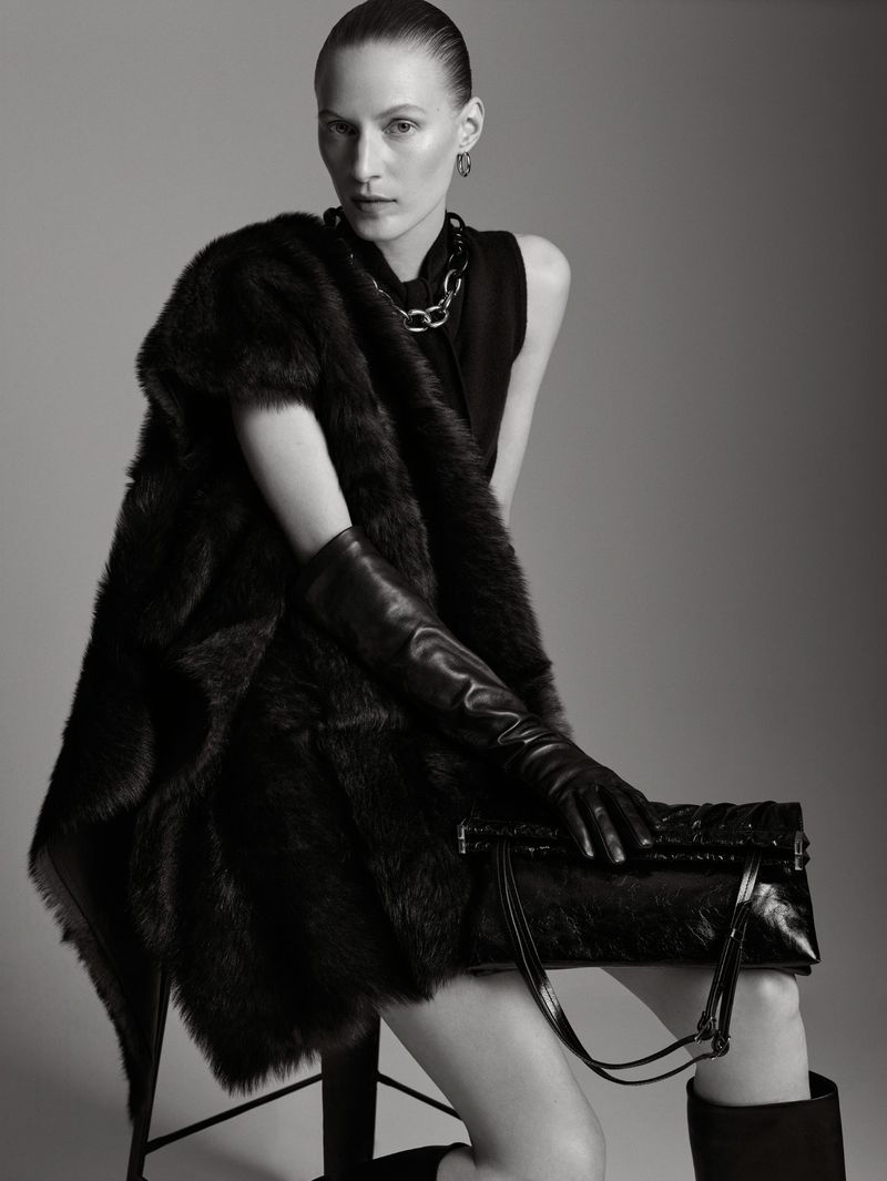 Elegant woman in black top, fur shawl, and leather gloves poses with a handbag in a studio setting, exuding high fashion.
