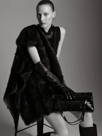 Elegant woman in black top, fur shawl, and leather gloves poses with a handbag in a studio setting, exuding high fashion.