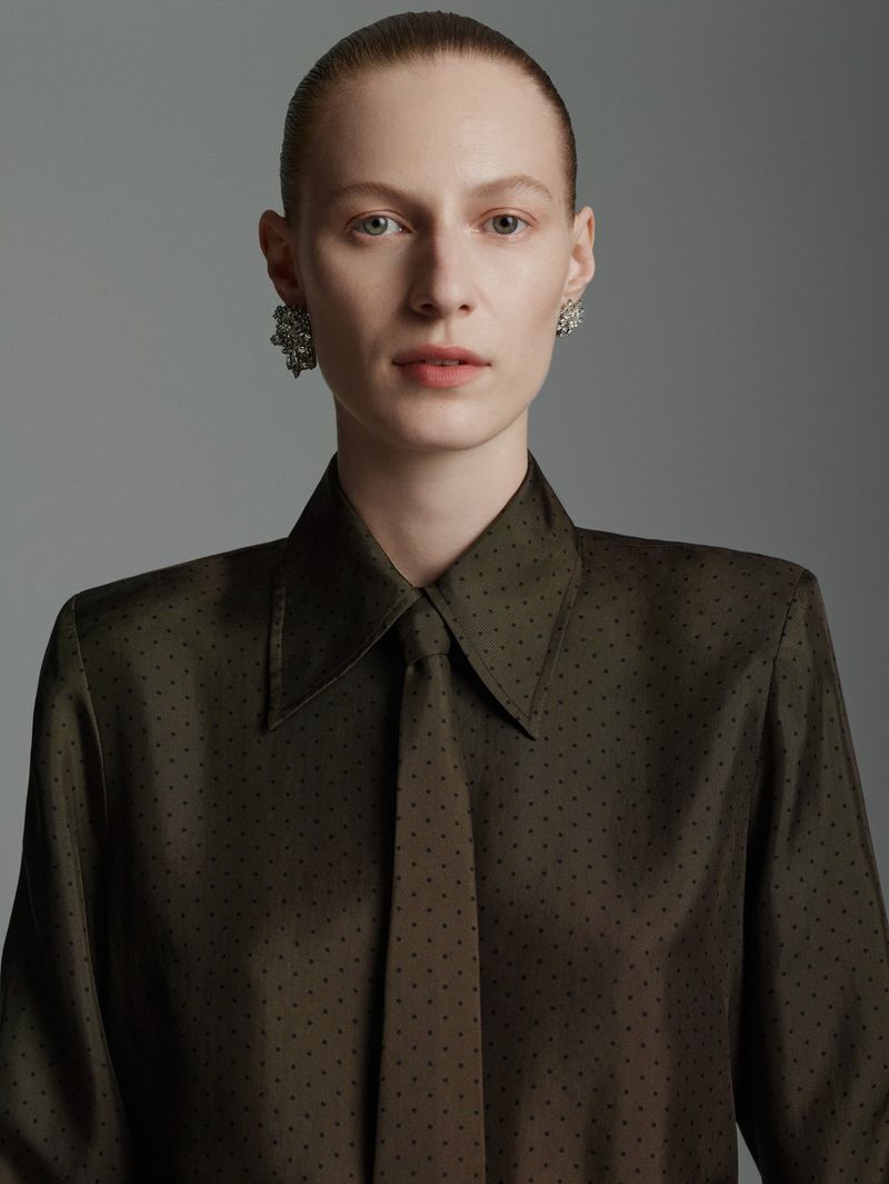 Close-up studio portrait of a fair-skinned adult female wearing an olive-green polka dot dress shirt and tie, with large jeweled earrings.