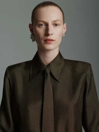 Close-up studio portrait of a fair-skinned adult female wearing an olive-green polka dot dress shirt and tie, with large jeweled earrings.