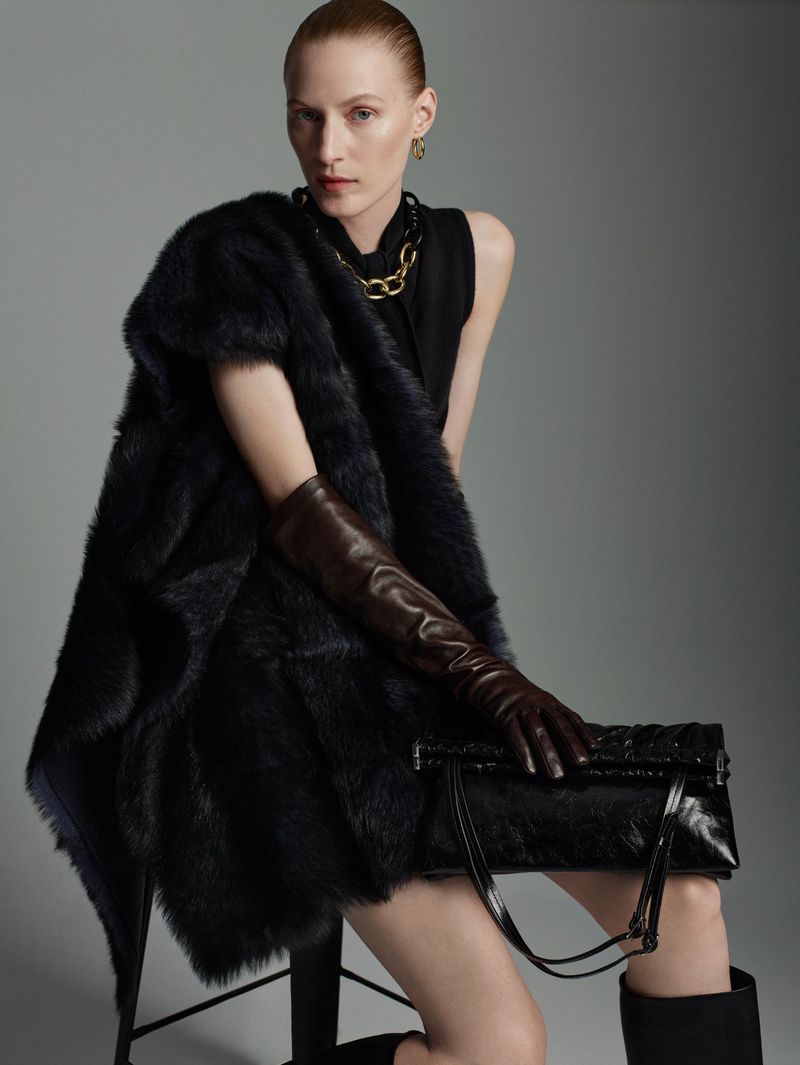 A red-haired woman is modeling while sitting, wearing a black dress, a black fur shawl, brown gloves, and a black purse against a gray background.