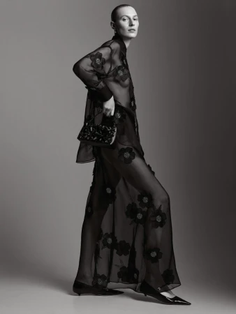 Model Julia Nobis wearing a black sheer floral blouse and skirt holding a handbag against a grey studio backdrop