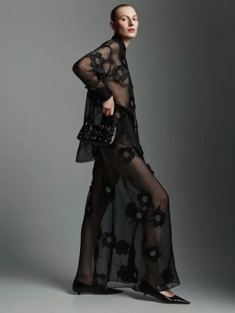 Model Julia Nobis wearing a black sheer floral shirt and matching skirt holding a small handbag in a studio
