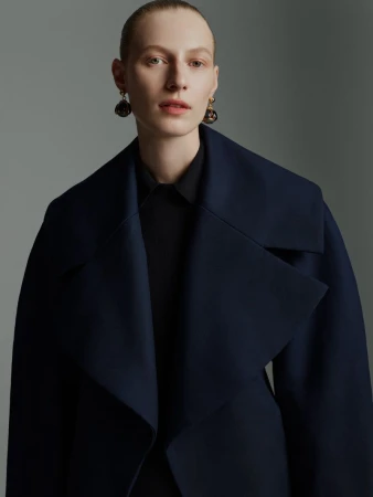 Model Julia Nobis wearing a dark structured coat and statement earrings against a neutral grey studio background.