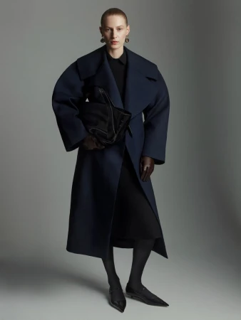 Model Julia Nobis wearing an oversized navy coat and holding a black handbag against a grey backdrop for Scanlan Theodore