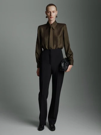 Model wearing a dark green silk shirt and black trousers holding a black handbag against a grey studio backdrop.