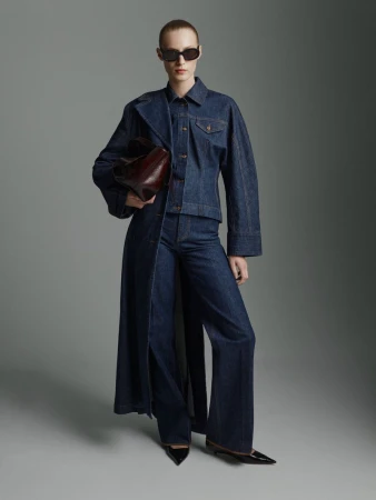Model Julia Nobis wearing a dark denim trench coat and carrying a dark red leather handbag against a grey seamless background.