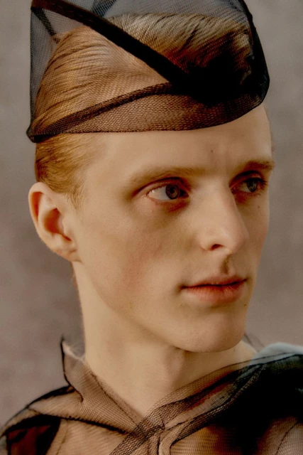 Close-up portrait of a male model with red hair wearing a black net hat and a translucent net top against a neutral grey backdrop.