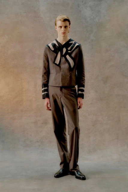 Full-length studio portrait of a young adult male in a brown sailor-style suit with black accents posing against a mottled brown backdrop.