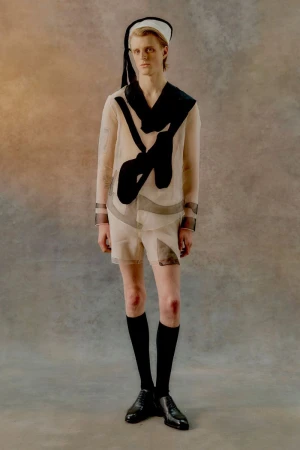 Young adult man stands in a sheer sailor suit with a black scarf and hat, black knee socks, and dress shoes against a mottled grey background.