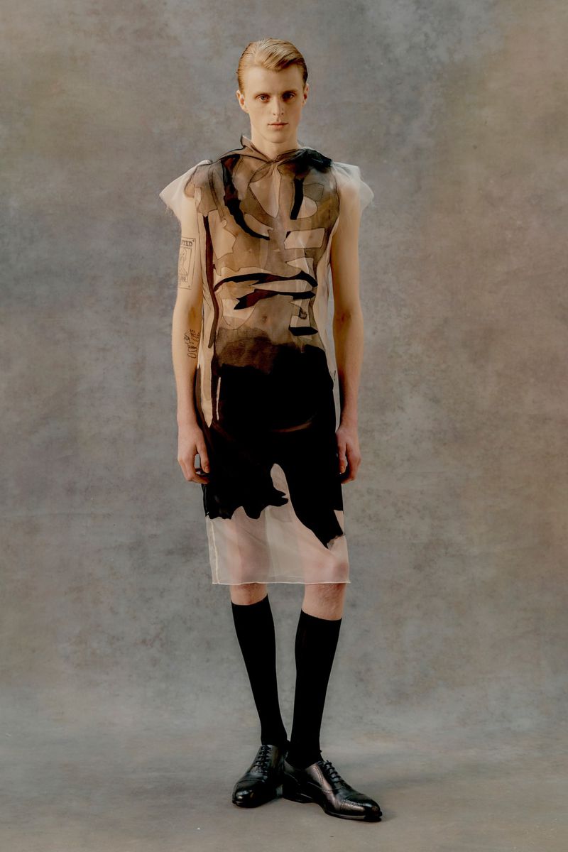 Fashion portrait of a blond male model in a sheer, sleeveless, graphic-print dress, black socks, and dress shoes on a mottled grey backdrop.