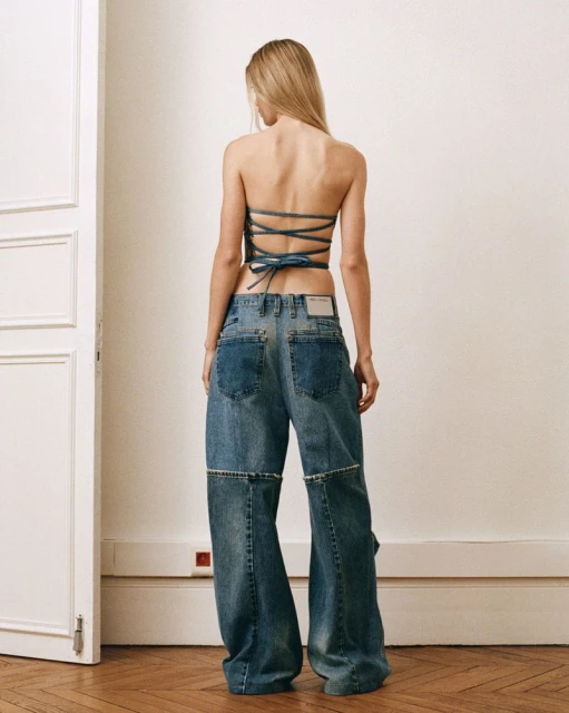 Blonde woman wearing a blue denim halter top and oversized jeans in an indoor setting, posing with her back to the camera.