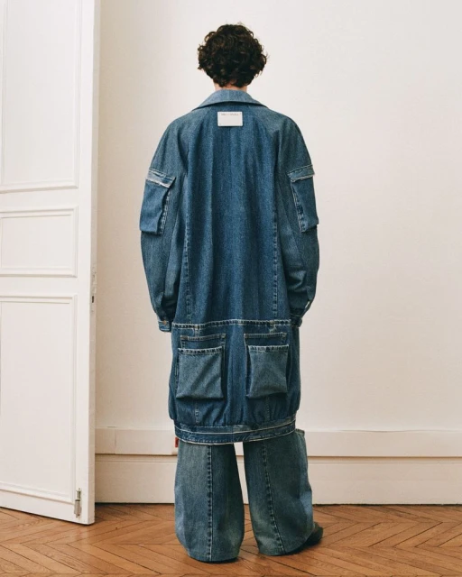 Person with curly hair wearing an oversized denim coat and wide leg pants poses in front of a white wall and a partially open white door.