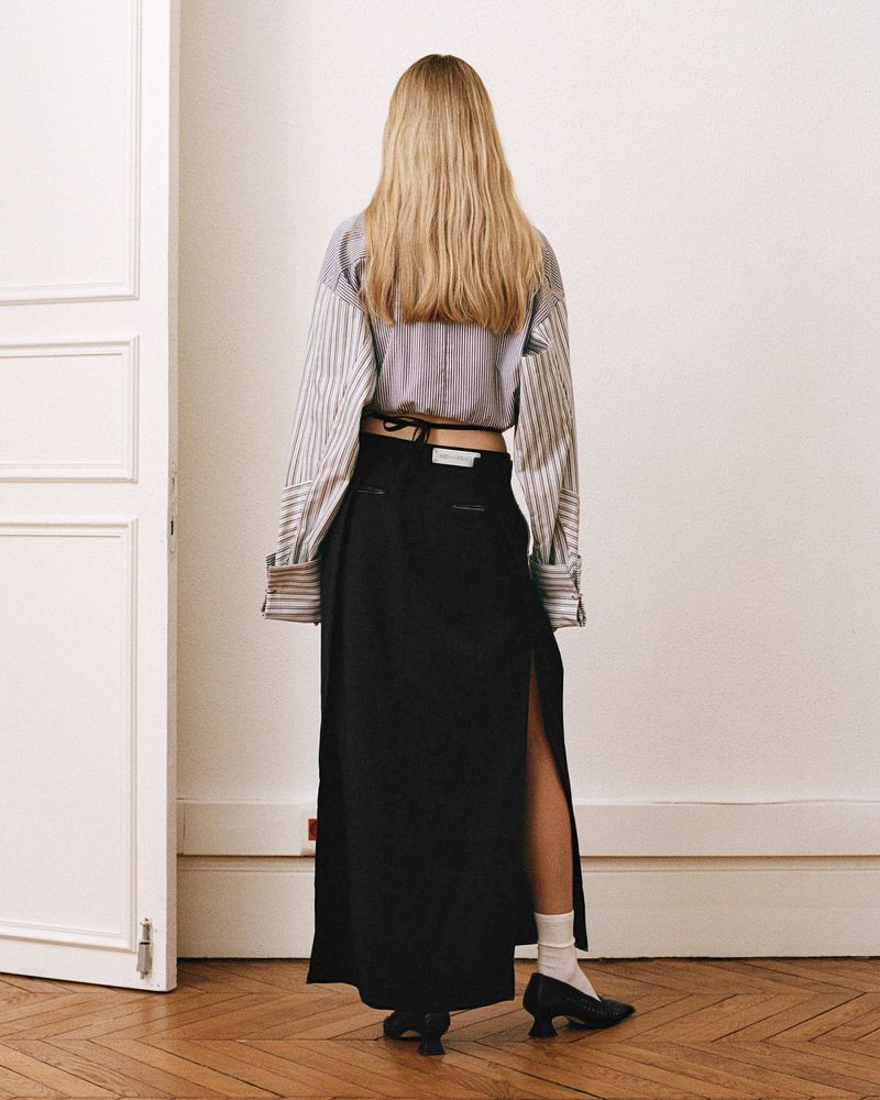 Back view of a blonde woman in a striped blouse and black maxi skirt with a slit standing in an indoor setting against a white wall.