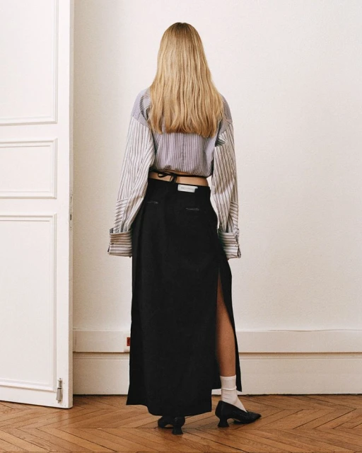 Back view of a blonde woman in a striped blouse and black maxi skirt with a slit standing in an indoor setting against a white wall.