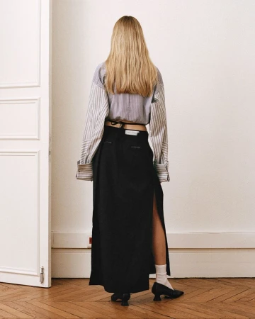 Back view of a blonde woman in a striped blouse and black maxi skirt with a slit standing in an indoor setting against a white wall.