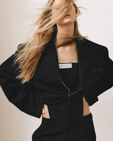 A fashionable blonde woman poses indoors wearing a black blazer, top, and pants, styled in a studio-like setting with neutral background.