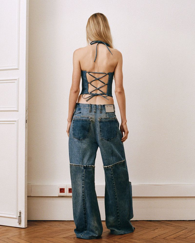 Back view of a woman in a denim halter top with lace-up back and baggy jeans, posing against a white wall with a partially open white door.