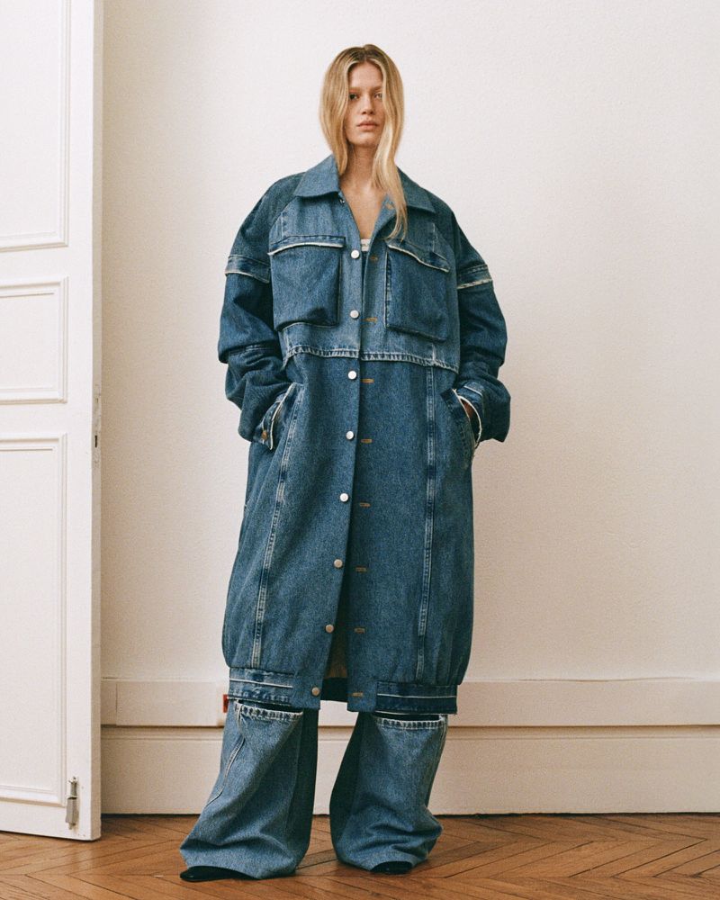 A woman stands in a light room modeling an oversized denim coat and matching wide-leg pants, showcasing a unique fashion statement.