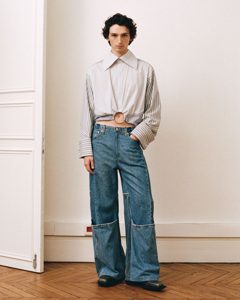 Adult male fashion model stands indoors wearing a striped crop-top shirt and wide-leg denim pants with knee cutouts. Light neutral walls.