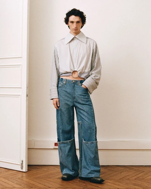 Adult male fashion model stands indoors wearing a striped crop-top shirt and wide-leg denim pants with knee cutouts. Light neutral walls.