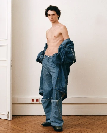 A slender adult male stands indoors, wearing oversized denim pants and jacket, shirtless, against a neutral wall with parquet flooring.