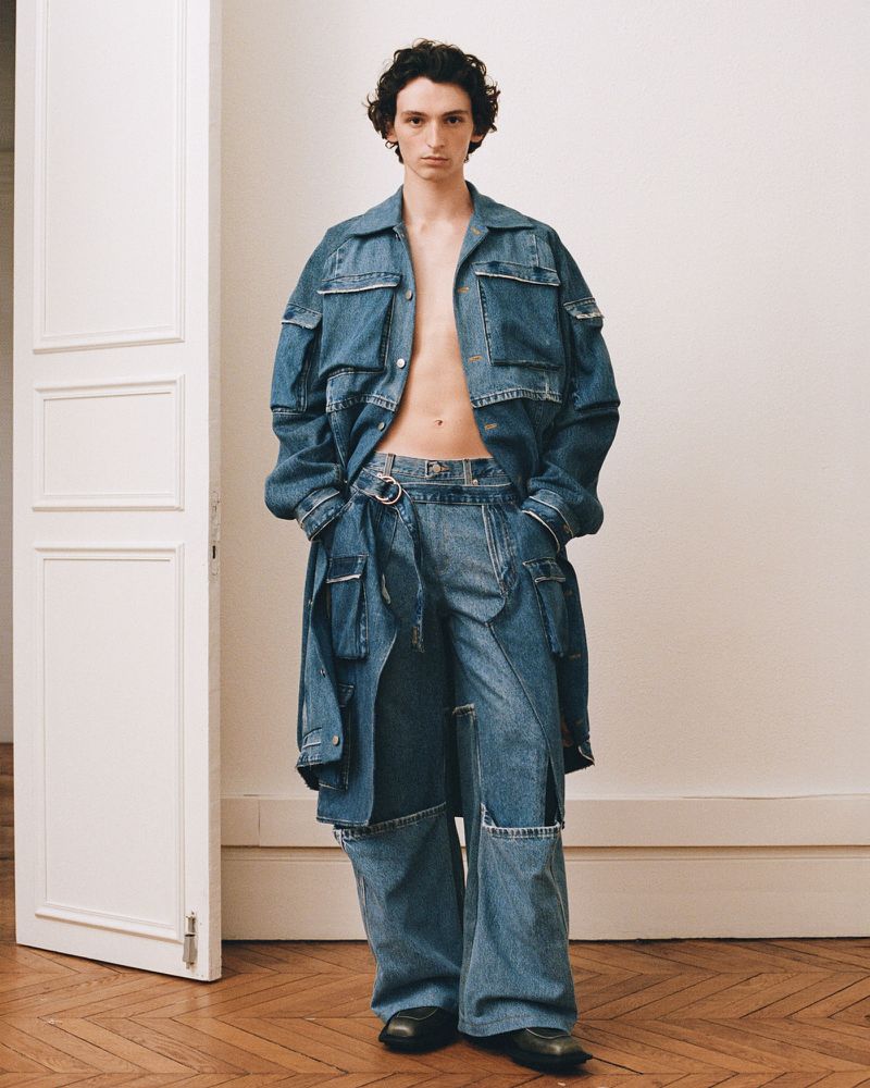 A male model wearing a denim utility jacket and matching trousers standing in an interior room with a paneled white door.