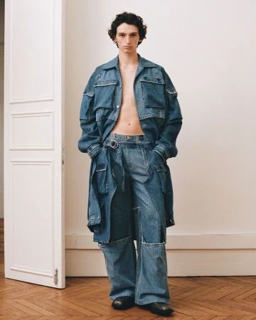 A male model wearing a denim utility jacket and matching trousers standing in an interior room with a paneled white door.