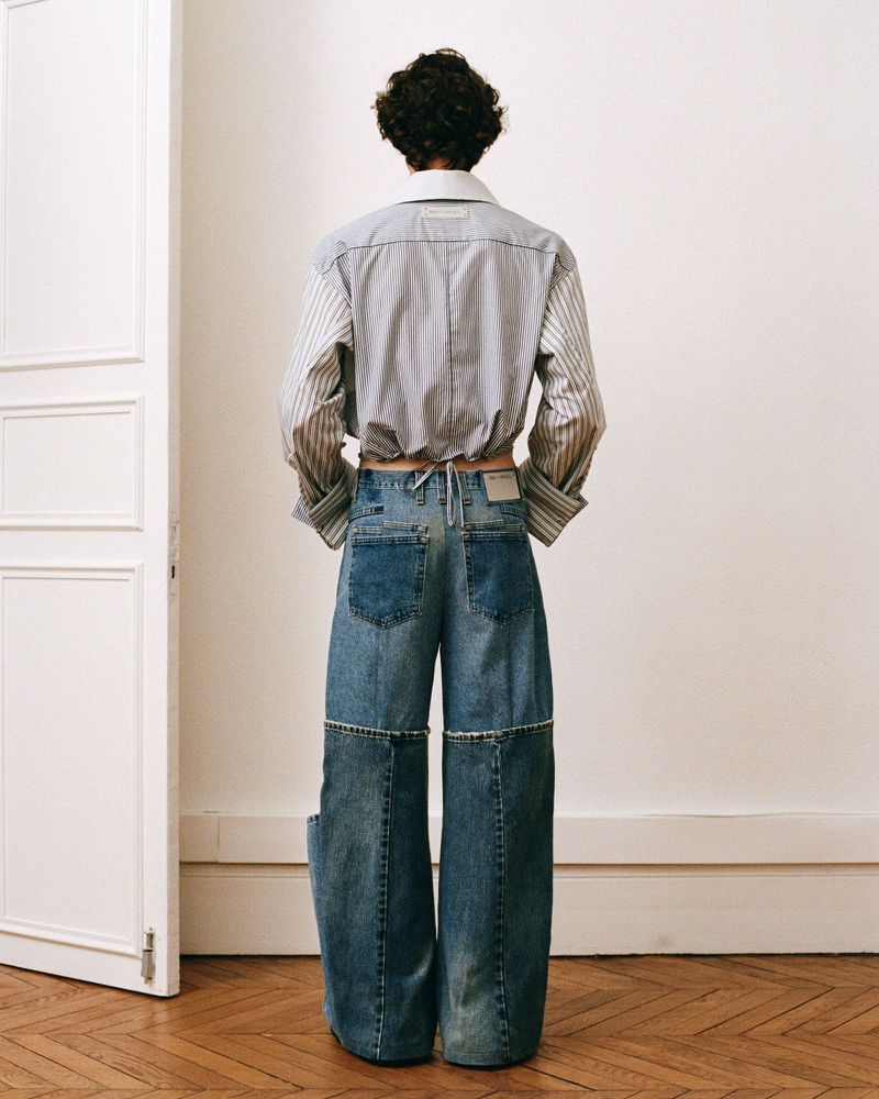 Rear view of a person wearing a striped button-down shirt tied at the waist and wide-leg denim pants in a studio with hardwood floors.