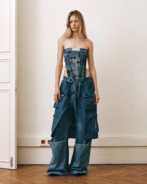 Woman in denim corset and wide-leg jeans stands in a light studio with wood floors. Fashion editorial shot. 