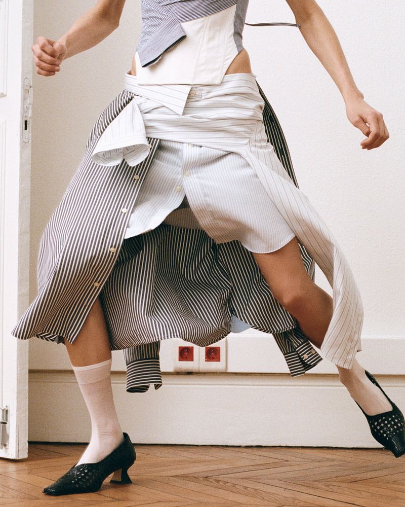 A woman in motion, wearing a deconstructed shirt dress, white socks, and black heels, standing on hardwood floor in front of a white door.