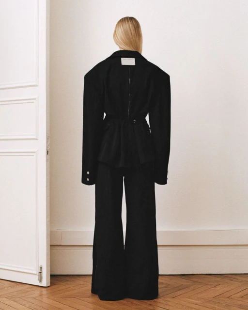 Fashion shot from behind of an adult female wearing a black blazer and pants, posing indoors with a white door visible on the left side.