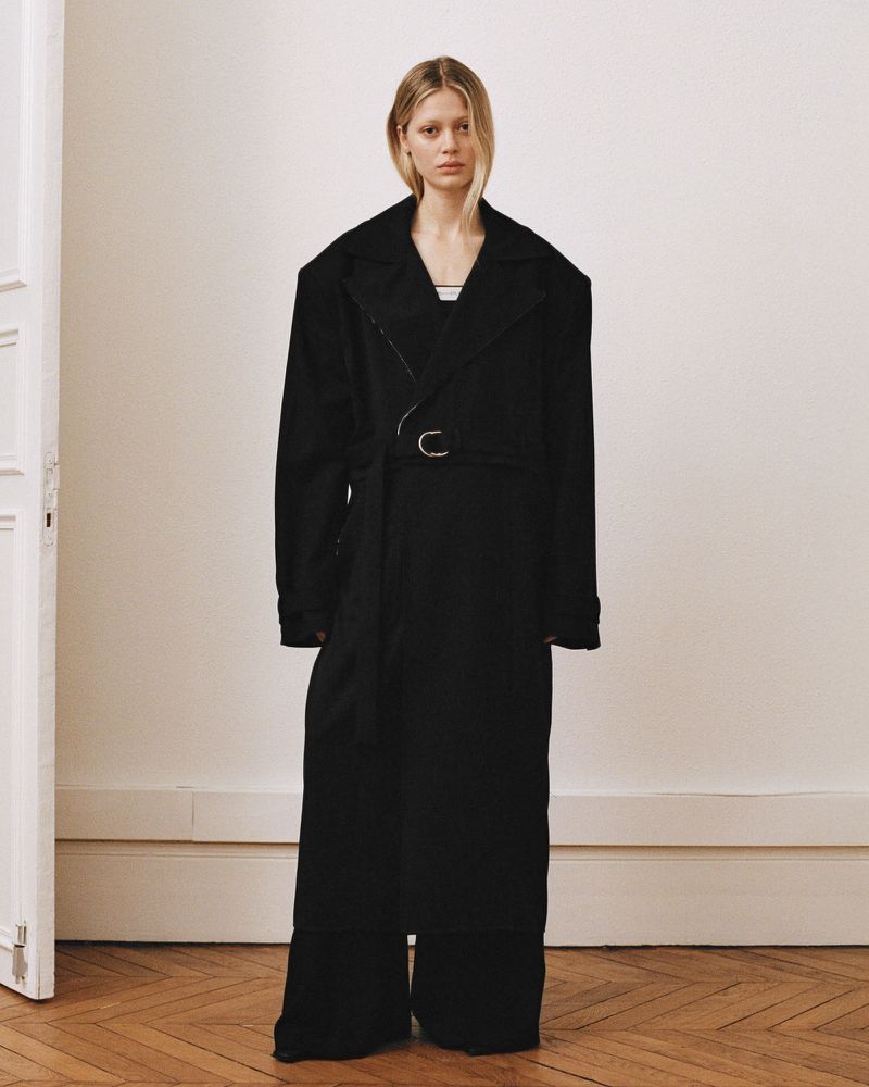 A slender adult woman poses indoors in a dramatically oversized black trench coat and matching pants, standing against an off-white wall.