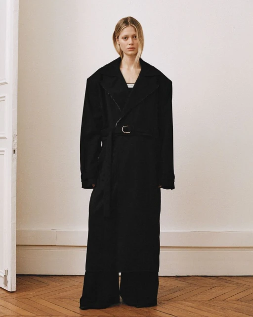 A slender adult woman poses indoors in a dramatically oversized black trench coat and matching pants, standing against an off-white wall.