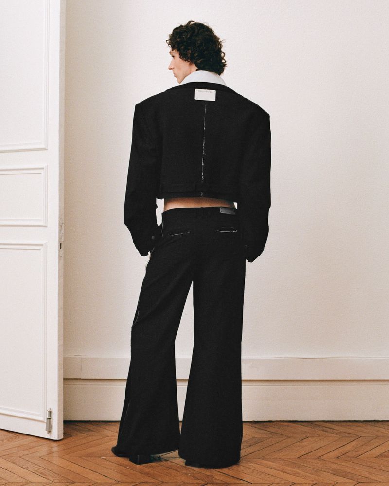 Back view of androgynous adult in black crop jacket and bell-bottom pants standing in a neutral indoor setting, fashion-forward.
