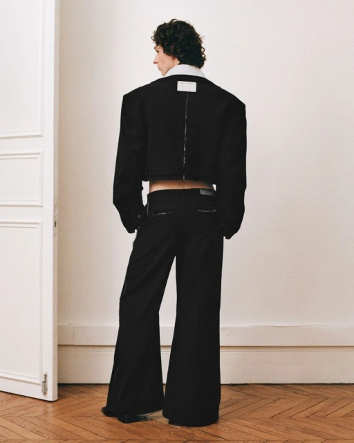 Back view of androgynous adult in black crop jacket and bell-bottom pants standing in a neutral indoor setting, fashion-forward.