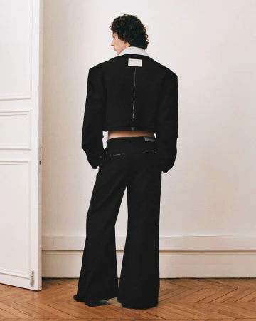 Back view of androgynous adult in black crop jacket and bell-bottom pants standing in a neutral indoor setting, fashion-forward.