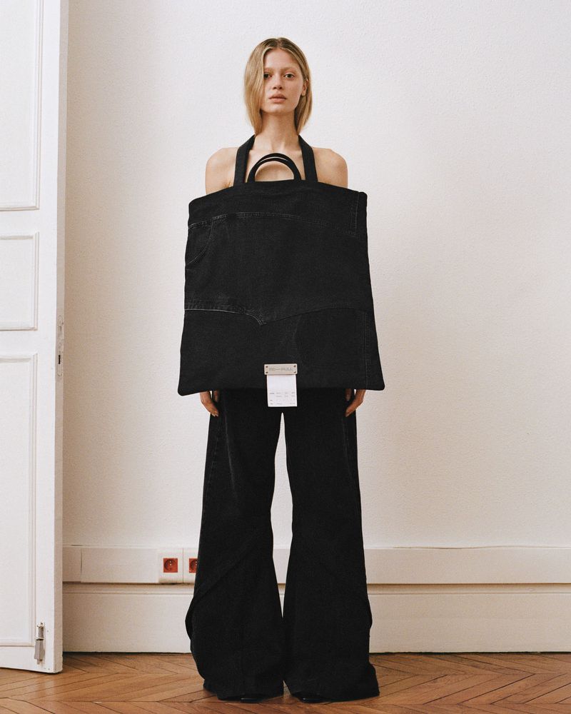Model standing indoors holding a large dark tote bag wearing black pants against a white wall.