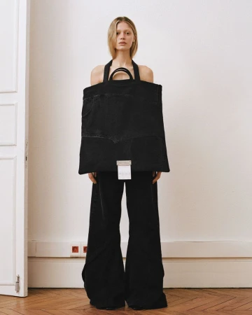 Model standing indoors holding a large dark tote bag wearing black pants against a white wall.