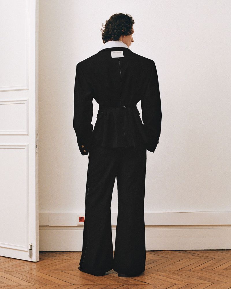 Back view of model in oversized black blazer and flared pants against a white wall with hardwood floor. Fashion editorial shot.