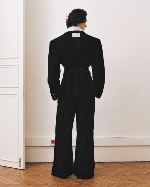 Back view of model in oversized black blazer and flared pants against a white wall with hardwood floor. Fashion editorial shot.