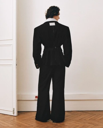 Back view of model in oversized black blazer and flared pants against a white wall with hardwood floor. Fashion editorial shot.