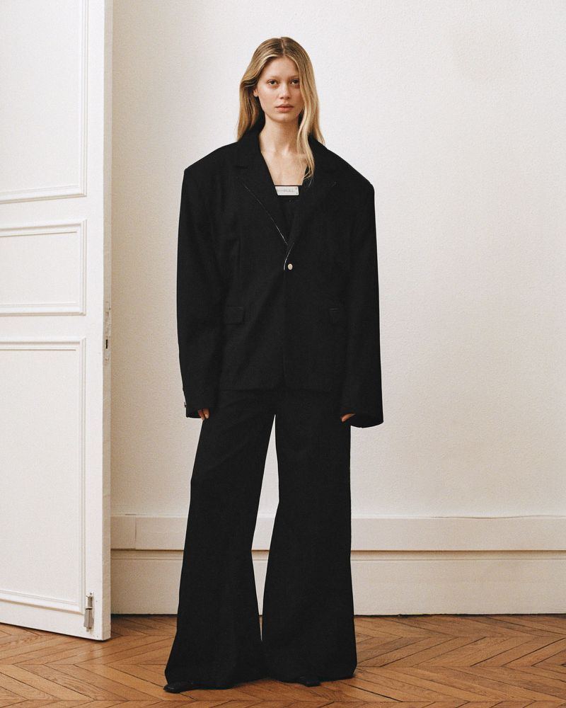 Model standing in a black oversized blazer and matching trousers against a pale wall with a partially open white door.