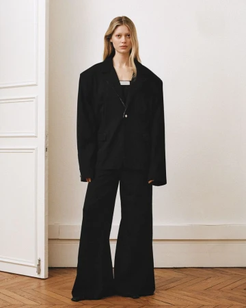Model standing in a black oversized blazer and matching trousers against a pale wall with a partially open white door.