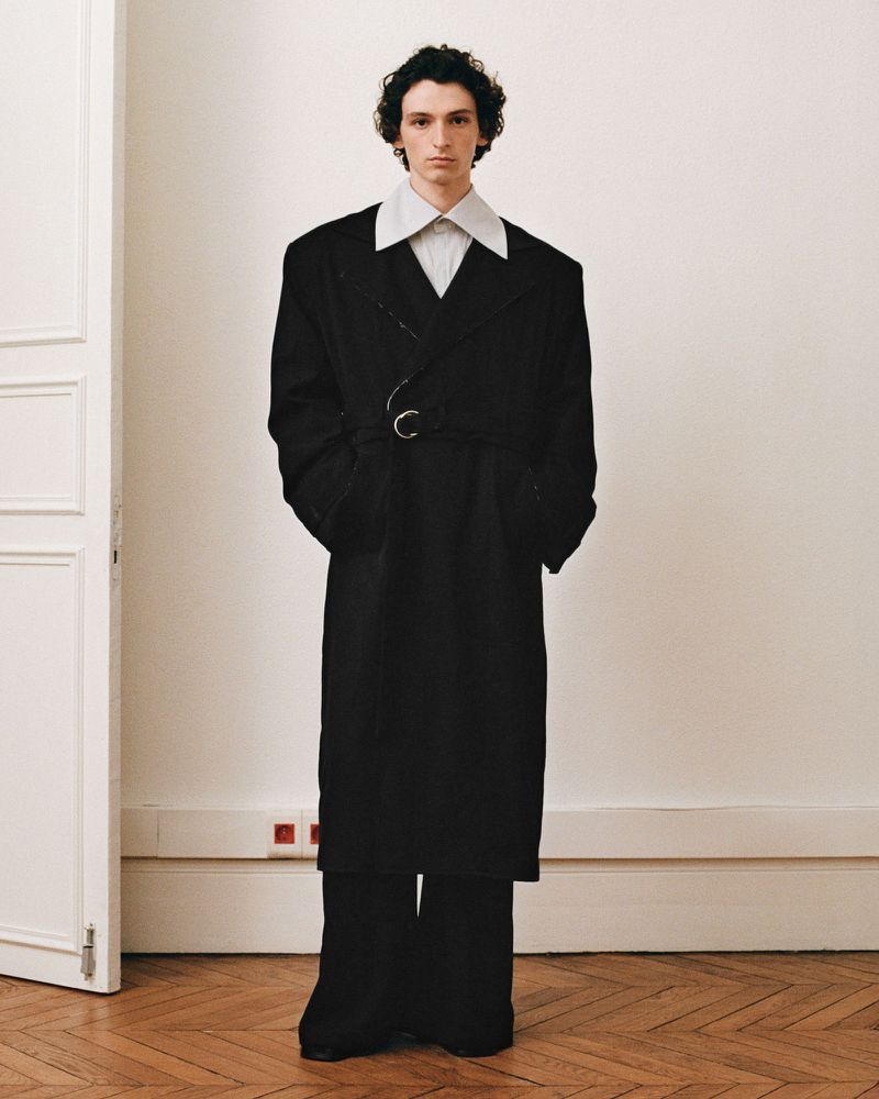 An adult male in an oversized black coat and wide-leg pants poses in front of an off-white wall with a door. Fashion editorial.