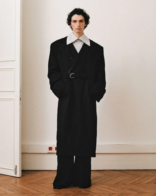 An adult male in an oversized black coat and wide-leg pants poses in front of an off-white wall with a door. Fashion editorial.