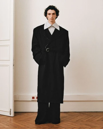 An adult male in an oversized black coat and wide-leg pants poses in front of an off-white wall with a door. Fashion editorial.
