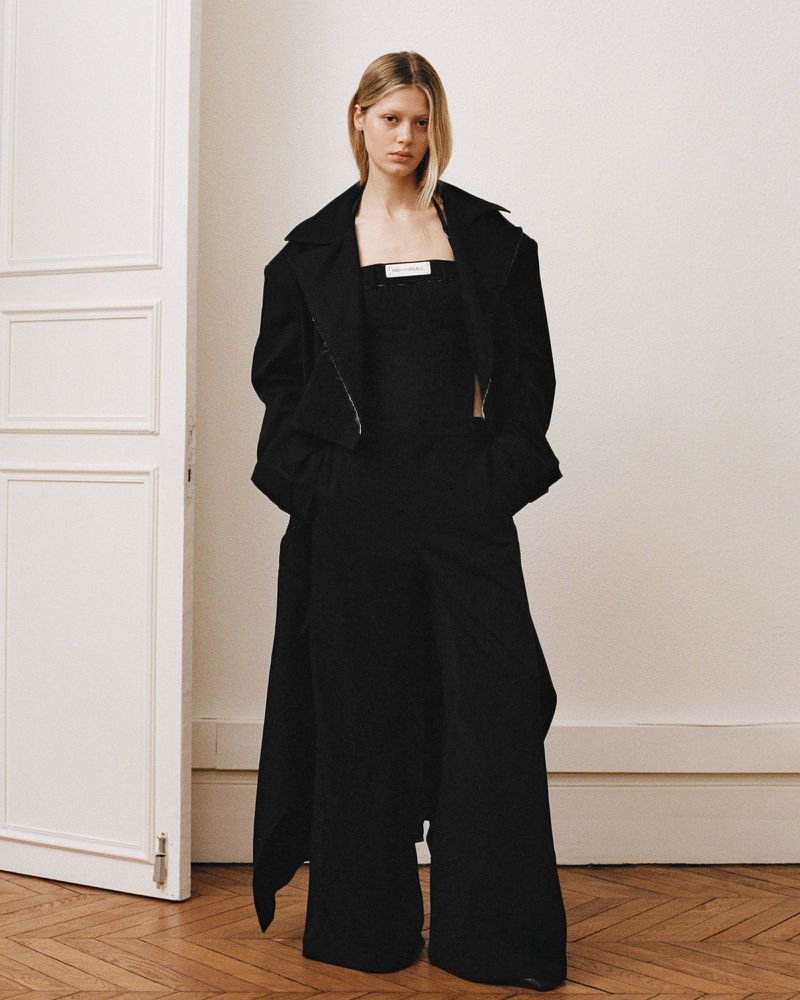 An adult female fashion model posing in black wide-leg pants, top and long coat in an indoor setting with hardwood flooring.