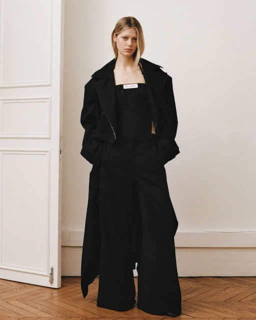 An adult female fashion model posing in black wide-leg pants, top and long coat in an indoor setting with hardwood flooring.