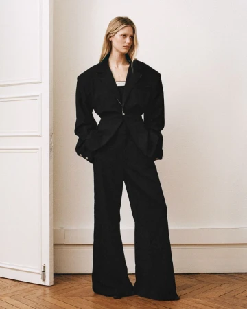 Model wearing a structured black blazer and wide leg trousers standing against a white wall with a partial door frame.